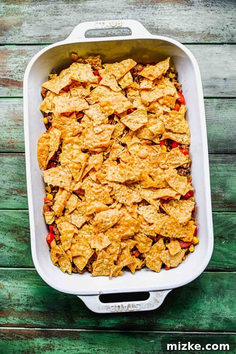 Adding a layer of crushed tortilla chips for crunch in the taco casserole. Roughly crushed tortilla chips are generously layered over the beef mixture in a white casserole dish, adding a crucial textural element to the taco casserole.