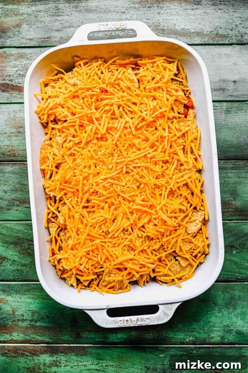 Topping the taco casserole with a generous layer of shredded cheese. A thick, even layer of shredded cheddar jack cheese covers the tortilla chips in a white casserole dish, ready to melt into a golden crust.