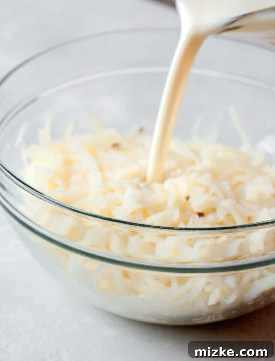 Liquid egg whites and heavy cream being meticulously poured into a large, clear mixing bowl that already contains a bed of shredded hashbrowns, preparing the base for the breakfast casserole.