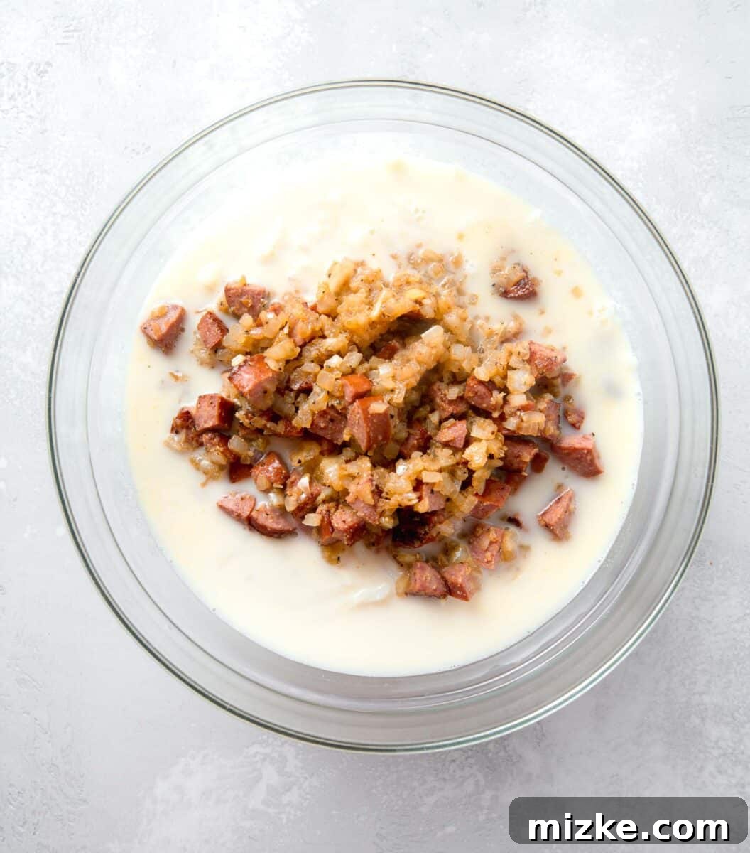 The cooled, savory sausage and onion mixture being carefully added to a large, clear mixing bowl, joining the combined liquid egg, cream, and shredded hashbrown mixture, ready for final seasoning and gentle stirring.
