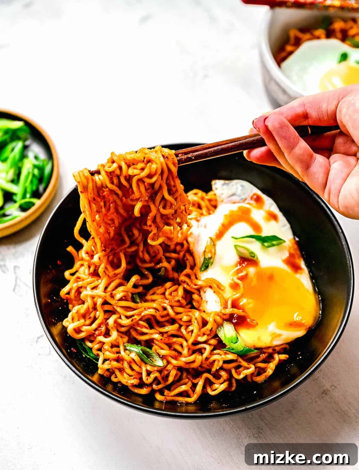 Instant ramen noodles are coated in a full-flavored, spicy sauce in this delicious, quick, and easy buldak ramen recipe. Chopsticks taking a bite of buldak ramen noodles, showcasing the rich, spicy sauce coating each strand.