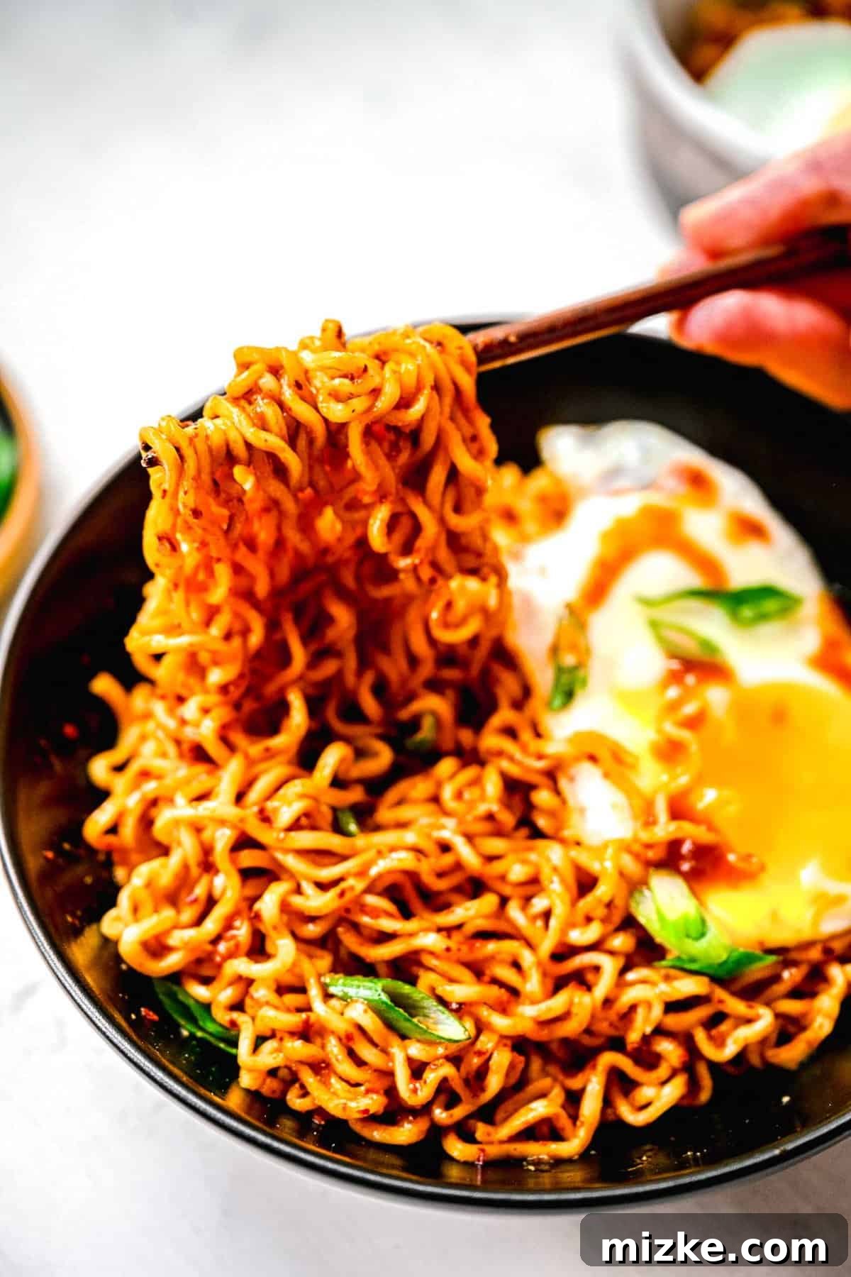 Instant ramen noodles are coated in a full-flavored, spicy sauce in this delicious, quick, and easy buldak ramen recipe. Chopsticks lifting a perfect bite of buldak ramen, showcasing the rich sauce and a hint of the runny yolk from the egg.