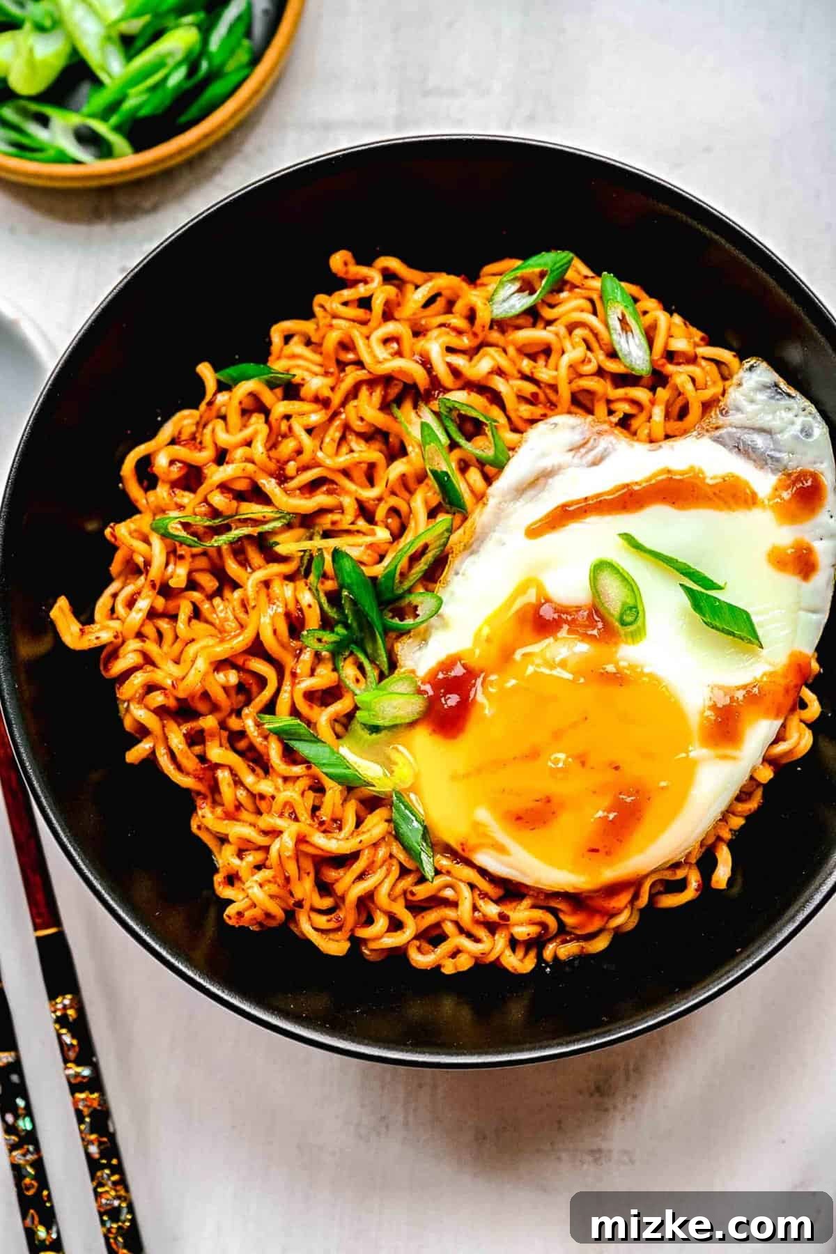 Instant ramen noodles are coated in a full-flavored, spicy sauce in this delicious, quick, and easy buldak ramen recipe. A steaming bowl of Buldak ramen, garnished with a sunny-side-up egg and fresh green onions, ready to be enjoyed.