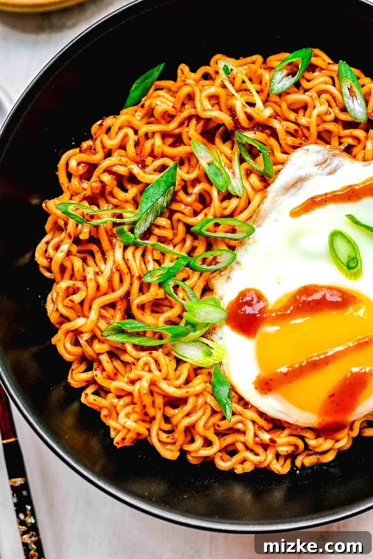 Instant ramen noodles are coated in a full-flavored, spicy sauce in this delicious, quick, and easy buldak ramen recipe. A close-up of Buldak ramen in a bowl, with chopsticks picking up a portion, highlighting the egg and sauce.