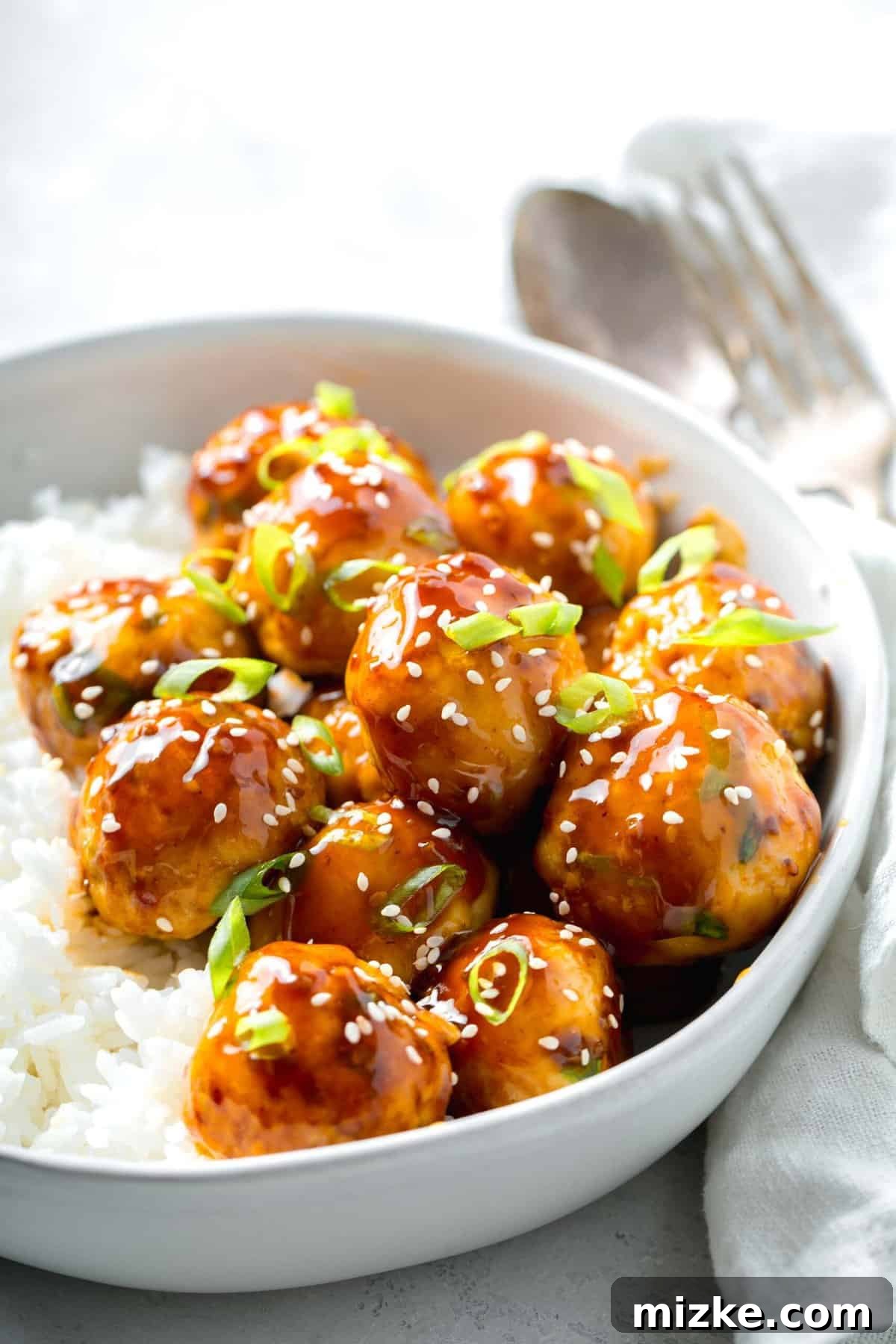 Juicy homemade teriyaki chicken meatballs, expertly blended with grated ginger, salty soy sauce, and other key seasonings for an explosion of flavor. Glazed teriyaki chicken meatballs garnished with fresh scallions and sesame seeds, served alongside fluffy white rice in a bowl, with a linen napkin nearby.