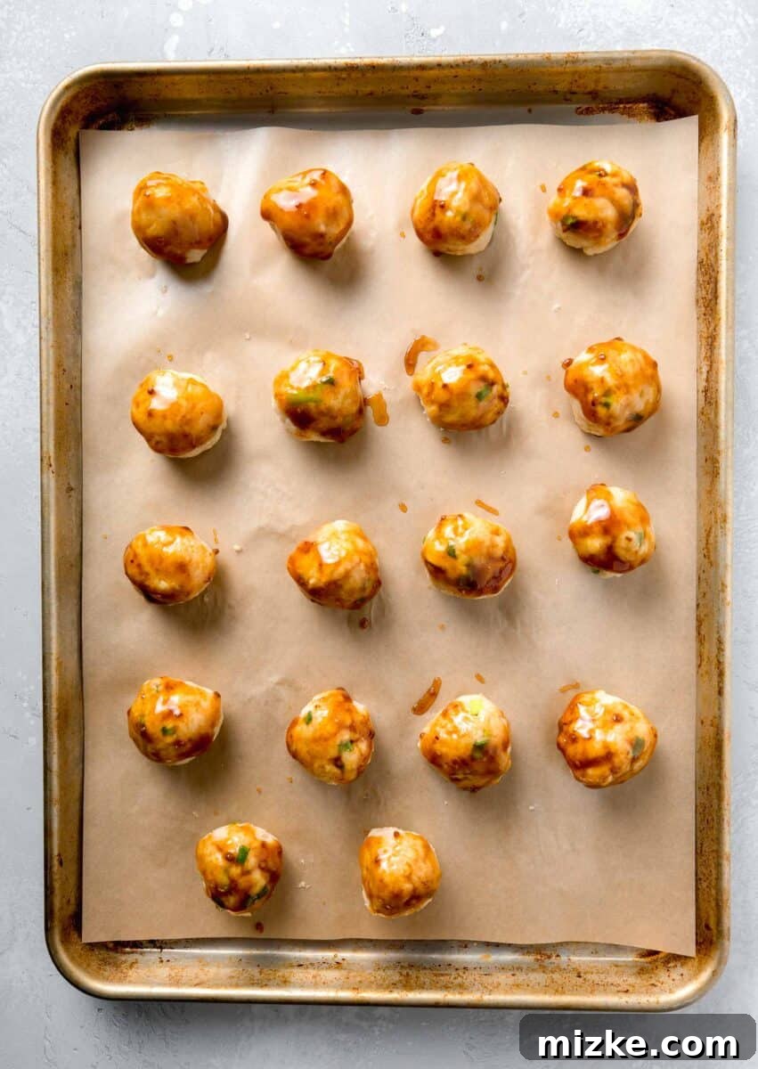 Raw ground chicken meatballs, enriched with grated ginger, salty soy sauce, and other seasonings, receiving their first glaze of teriyaki sauce before baking. Teriyaki chicken meatballs, freshly formed and lightly brushed with teriyaki sauce, meticulously arranged on parchment paper on a rimmed baking sheet.
