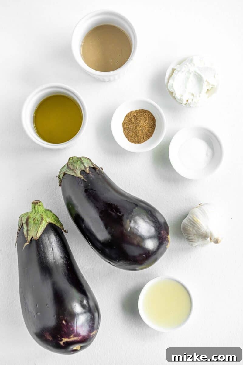 Smoky and Simple Baba Ganoush 3 Fresh ingredients for Baba Ganoush laid out on a white surface, including whole eggplants, a lemon, garlic cloves, and a bowl of tahini.