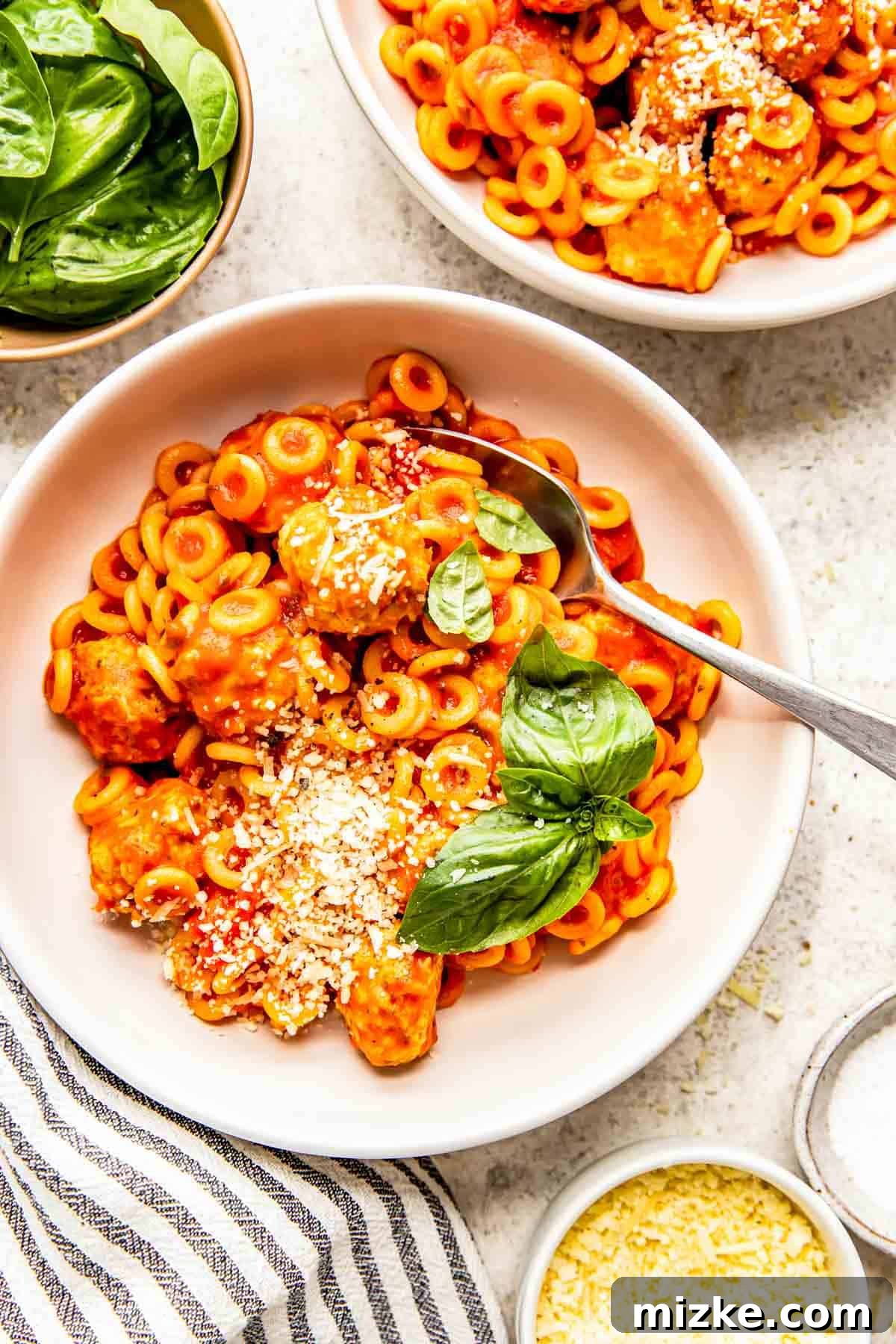 Homemade Tomato Pasta O's 3 A white bowl filled with homemade Spaghettios and chicken pesto meatballs, with a metal spoon inserted, topped with freshly grated Parmesan cheese and vibrant basil leaves.