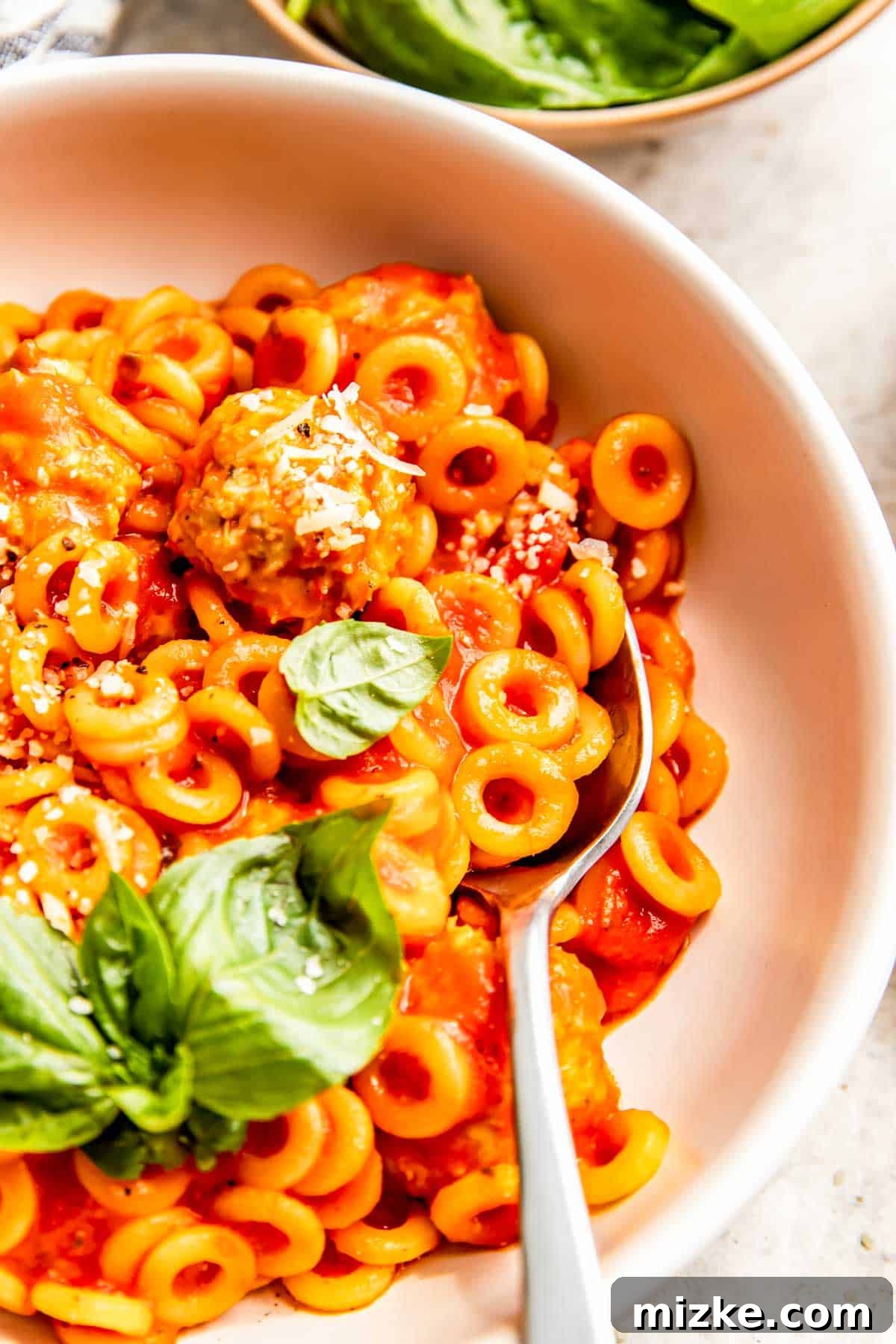 Homemade Tomato Pasta O's 4 A white bowl filled with homemade Spaghettios and chicken pesto meatballs, with a metal spoon, garnished with fresh basil leaves on the side.