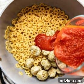 Rich marinara sauce being poured over cooked O-shaped pasta and chicken meatballs in a large pot, ready to be mixed.