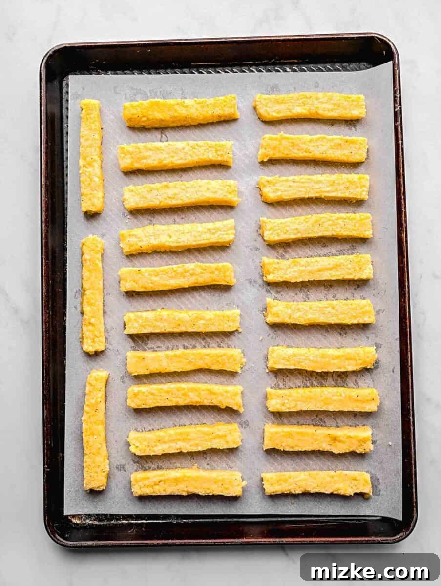 These polenta fries are made from cheesy polenta cut into strips, brushed with olive oil, and baked to crispy golden brown. Polenta strips on a baking sheet lined with parchment paper ready to be baked.