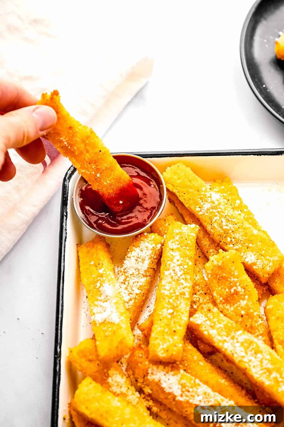 These polenta fries are made from cheesy polenta cut into strips, brushed with olive oil, and baked to crispy golden brown. Dipping a polenta fry in ketchup.
