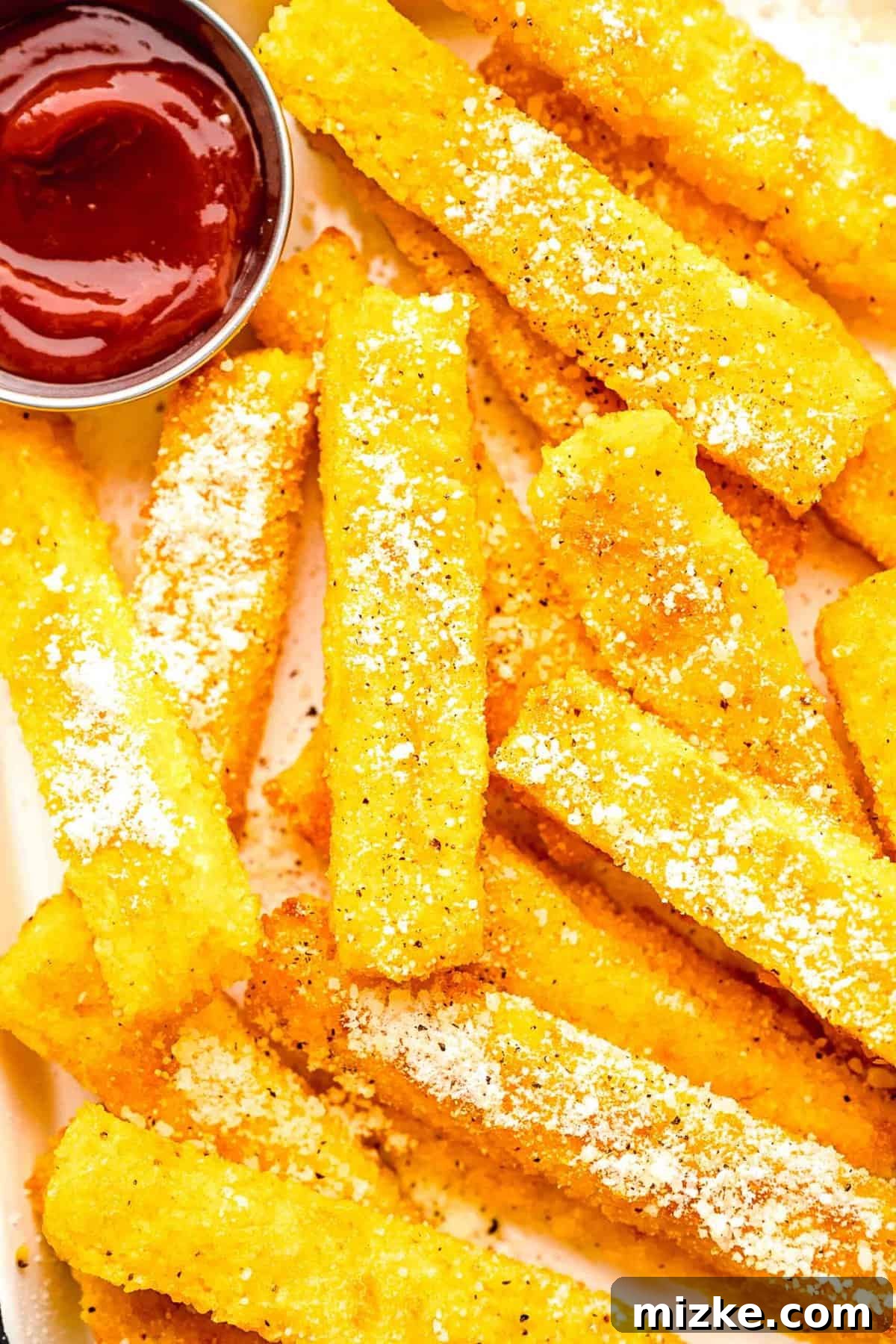 These polenta fries are made from cheesy polenta cut into strips, brushed with olive oil, and baked to crispy golden brown. Closeup of polenta fries with ketchup.