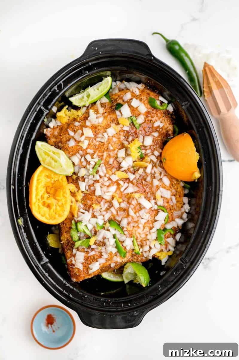 Melt-Away Slow Cooker Pork Carnitas 8 A whole jalapeño, sliced onion, and citrus halves are spread across the top of uncooked pork in a slow cooker.