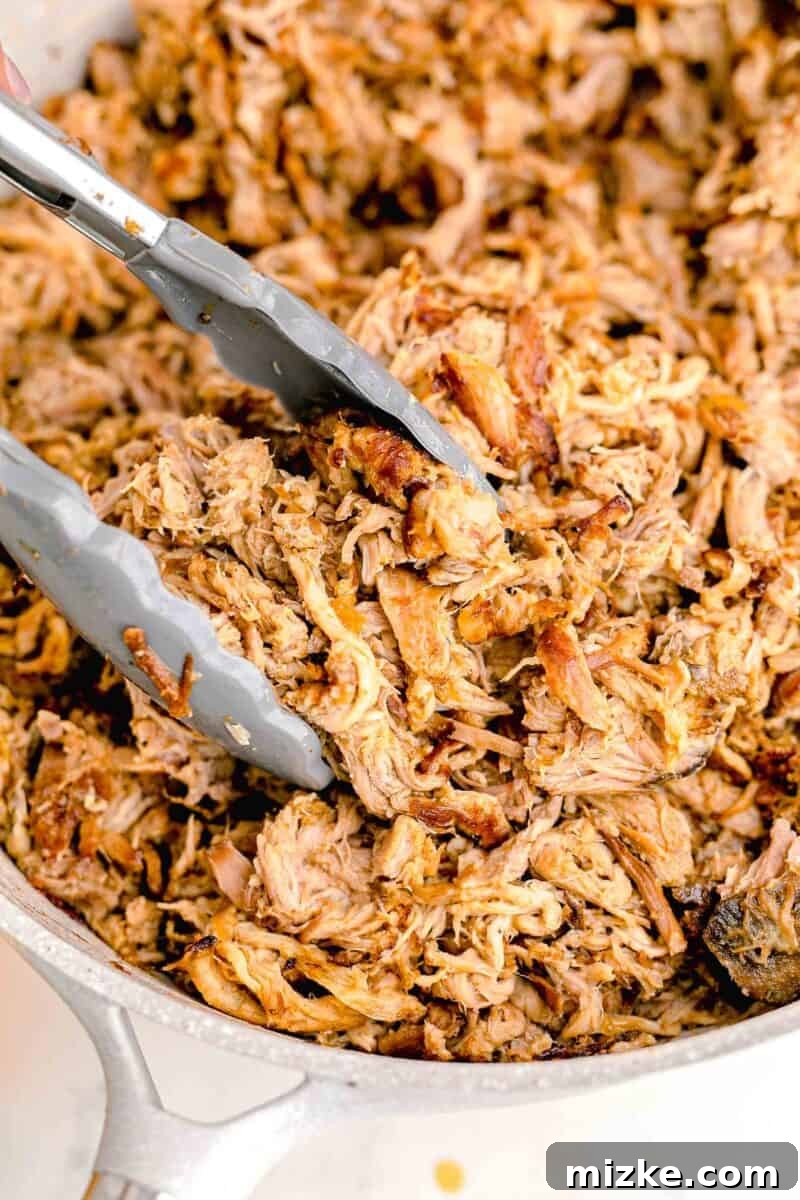Serving tongs are positioned in a rustic bowl filled with generous amounts of freshly cooked, shredded pork carnitas, ready to be served.