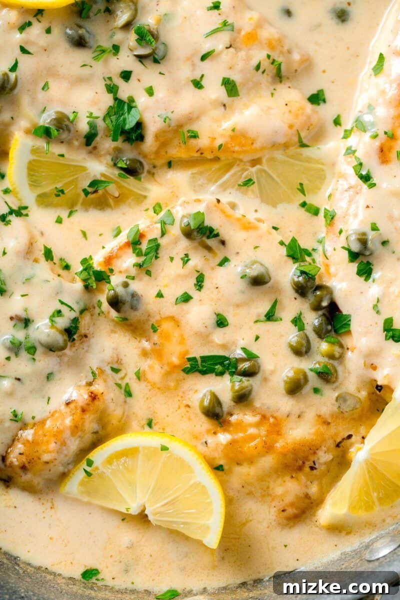 Velvety Lemon Chicken Piccata 10 A close-up of a serving of creamy lemon chicken piccata, featuring tender chicken breasts bathed in sauce, adorned with fresh parsley and capers, with a bright lemon wedge on the side.