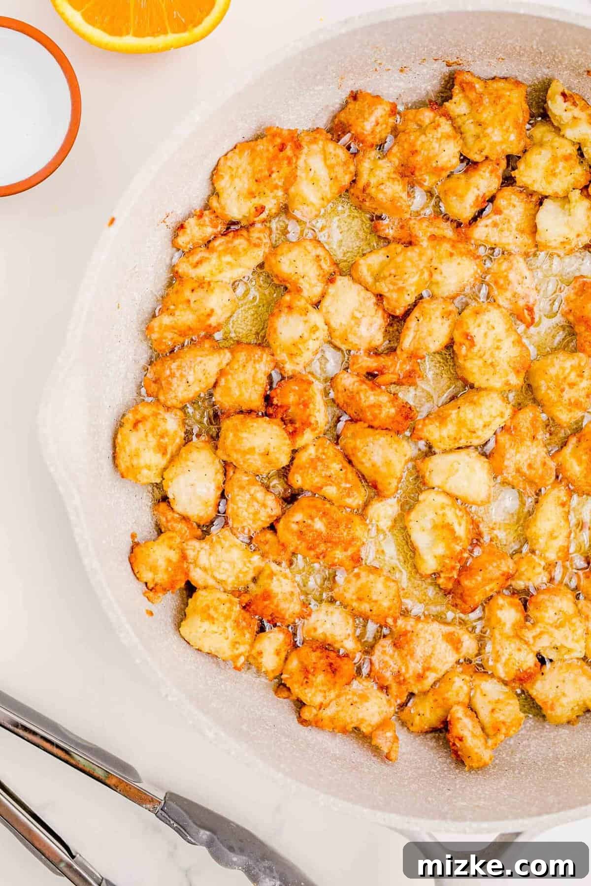 Achieving ideal crispiness on your homemade orange chicken. Close-up of golden-brown, crispy fried chicken bites in a skillet, nearly ready to be removed.