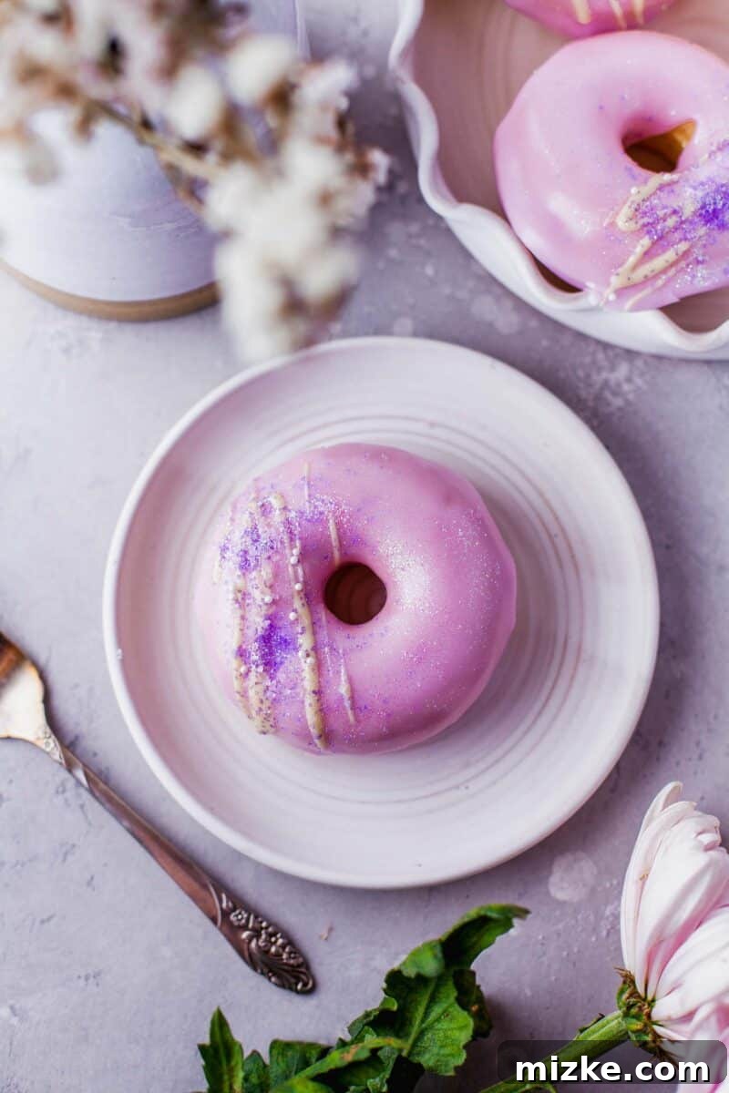 Perfectly Baked Vanilla Delights 8 A single beautifully frosted pink vanilla donut, adorned with a white chocolate drizzle, rests on a simple round white plate.