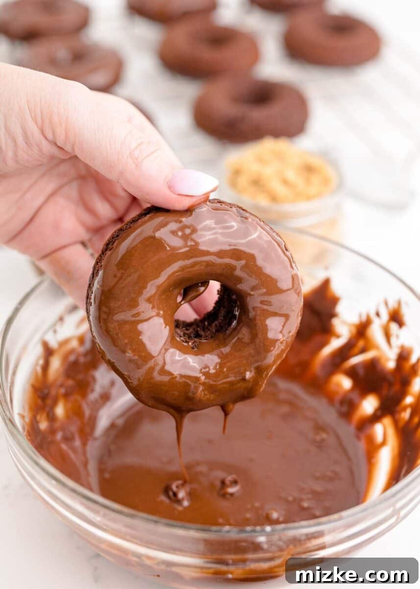 A baked donut being dipped into a bowl of rich chocolate frosting, ensuring a generous coating.