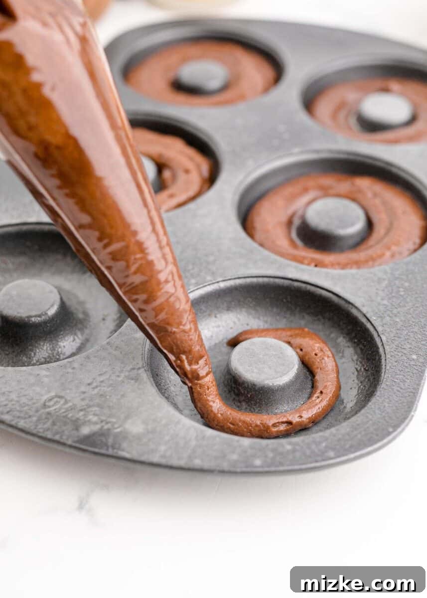 Chocolate donut batter being carefully piped into the prepared donut tins using a piping bag, ensuring even filling.