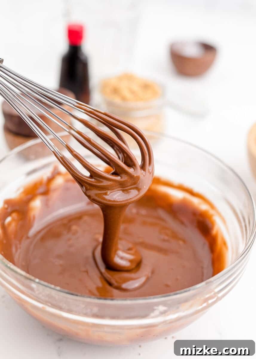 A whisk lifting and drizzling rich chocolate frosting over a bowl of more frosting, showing its smooth, glossy texture.