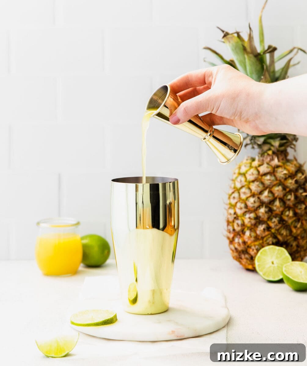 Golden pineapple juice is being poured into a sleek cocktail shaker filled with ice, ready to mix a refreshing drink.