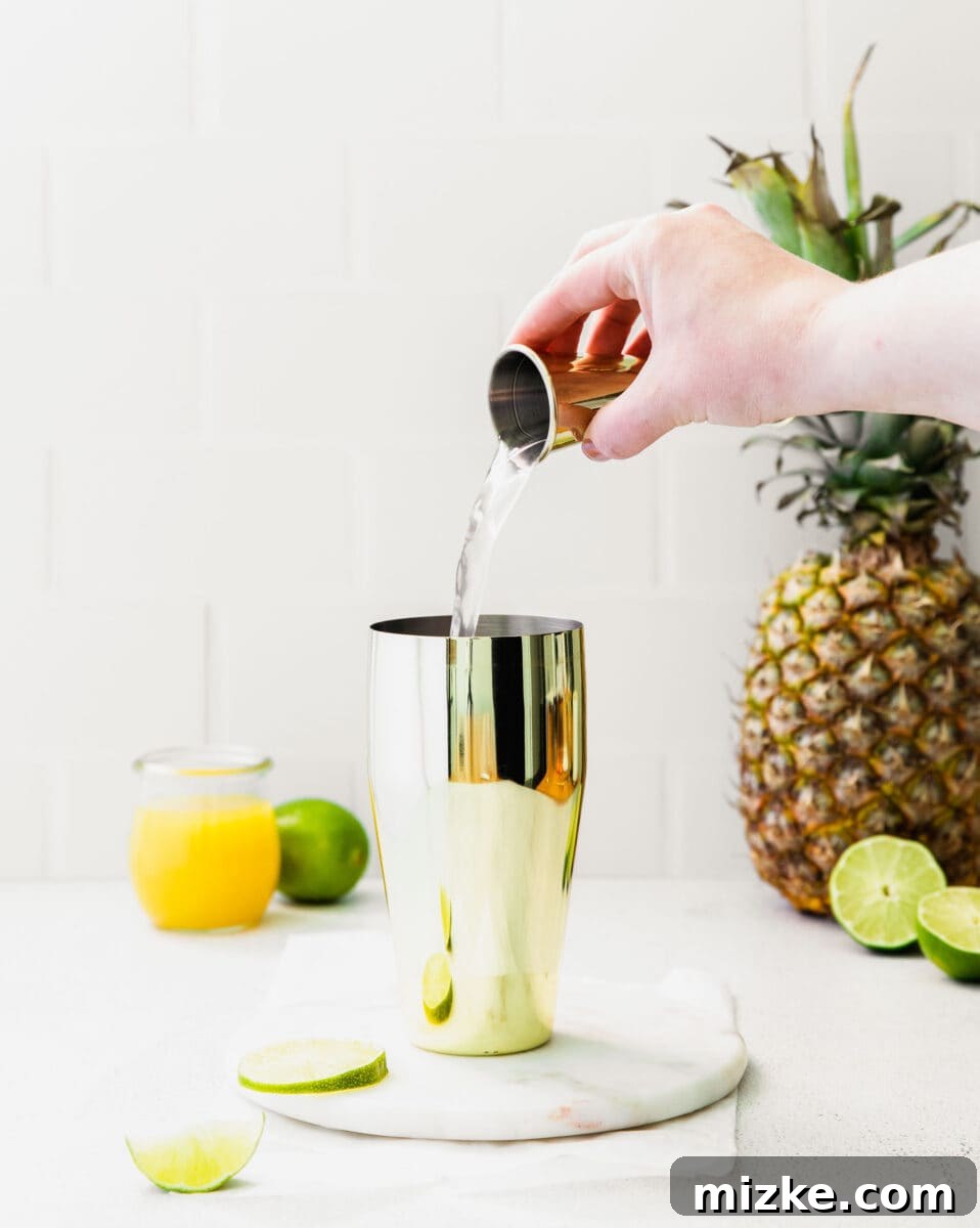 Clear liquid is carefully being poured from a small shot glass into a cocktail shaker, adding a measured ingredient to the mix.