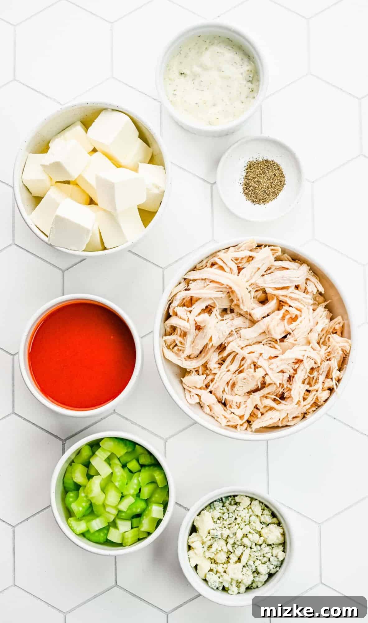 Crockpot buffalo chicken dip is the perfect party snack. It's creamy, loaded with chicken, and bursting with blue cheese and buffalo flavor. Various ingredients for crockpot buffalo chicken dip meticulously separated into small bowls, showcasing fresh celery, shredded chicken, cream cheese, blue cheese, and hot sauce.