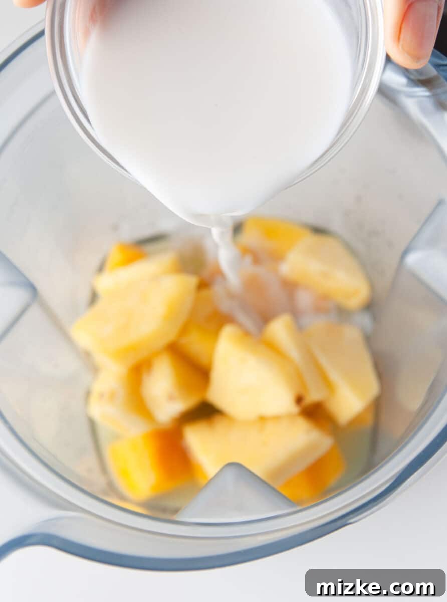 Velvet Mango Sunset 5 Rich coconut cream is being poured into a blender alongside the frozen mango chunks, creating the creamy base of the colada.