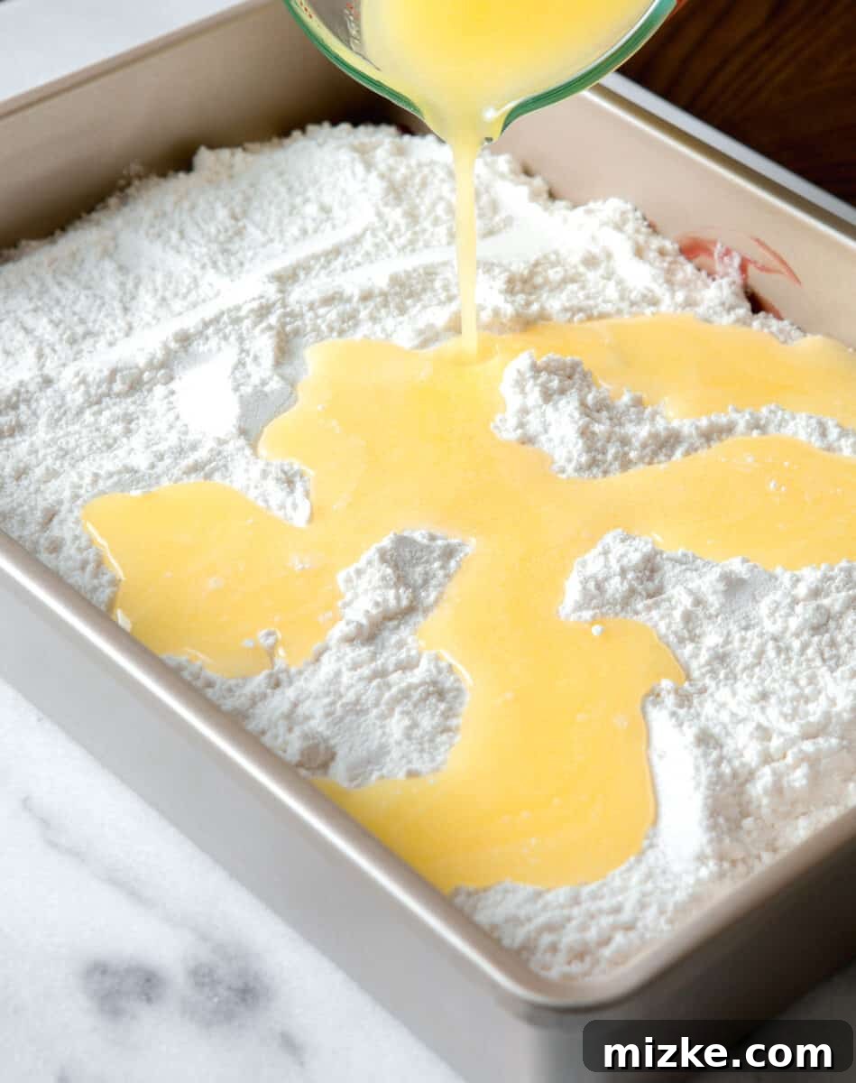 Effortless Cherry Delight 7 Melted butter being drizzled over the dry white cake mix layer in a metal baking pan, saturating the mix.