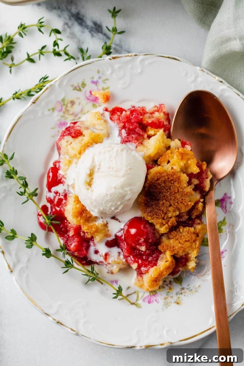 Effortless Cherry Delight 8 An overhead view of a scoop of vanilla ice cream melting on warm cherry dump cake, served on a floral dessert plate with a copper spoon and thyme sprigs.