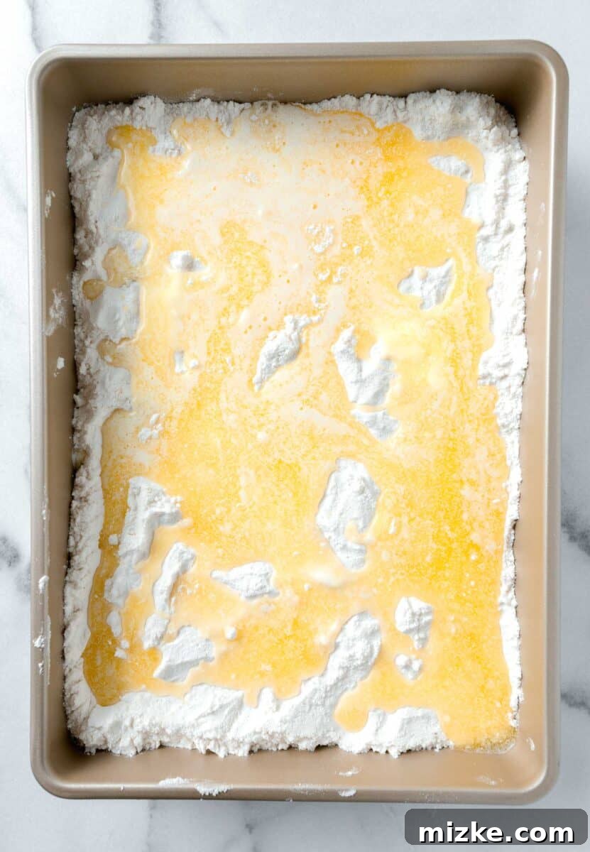 Melted butter poured generously over the layer of white cake mix in a 9x13 inch gold baking dish, ready for baking.