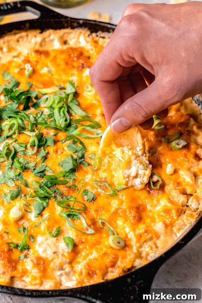 a hand holding a tortilla chip to scoop corn dip in a cast iron skillet