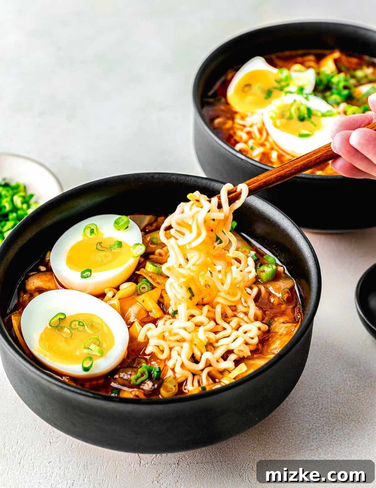 Enjoying Kimchi Ramen: A Quick and Flavorful Korean Noodle Soup Two bowls of kimchi ramen, with one hand holding chopsticks poised to enjoy a bite. The ramen is garnished with egg and green onions.