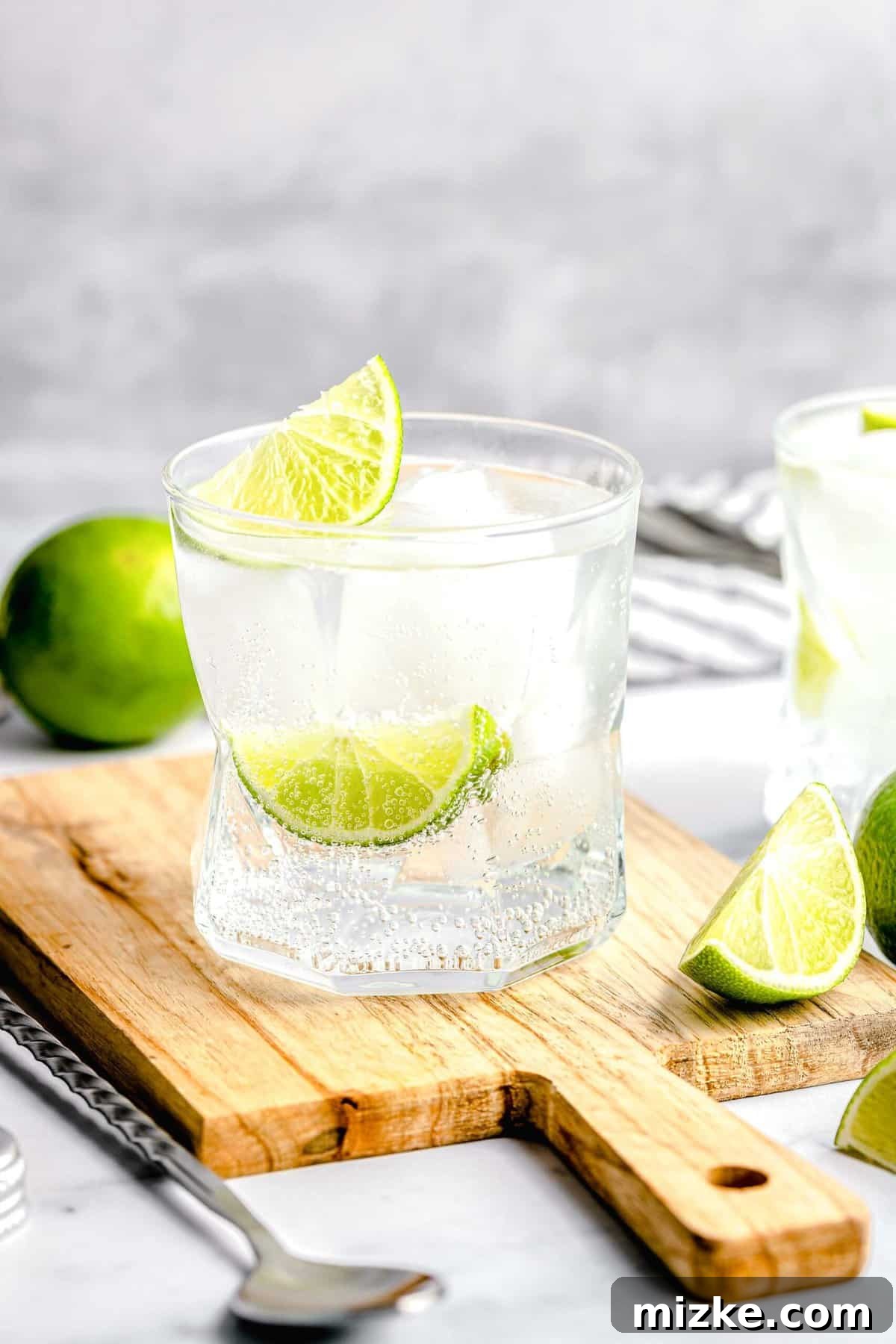 A fizzy, botanical gin and tonic, made with 1 part gin and 2 parts tonic, garnished with lime and ready to enjoy. Two gin and tonics on a cutting board, one in focus near a lime slice.