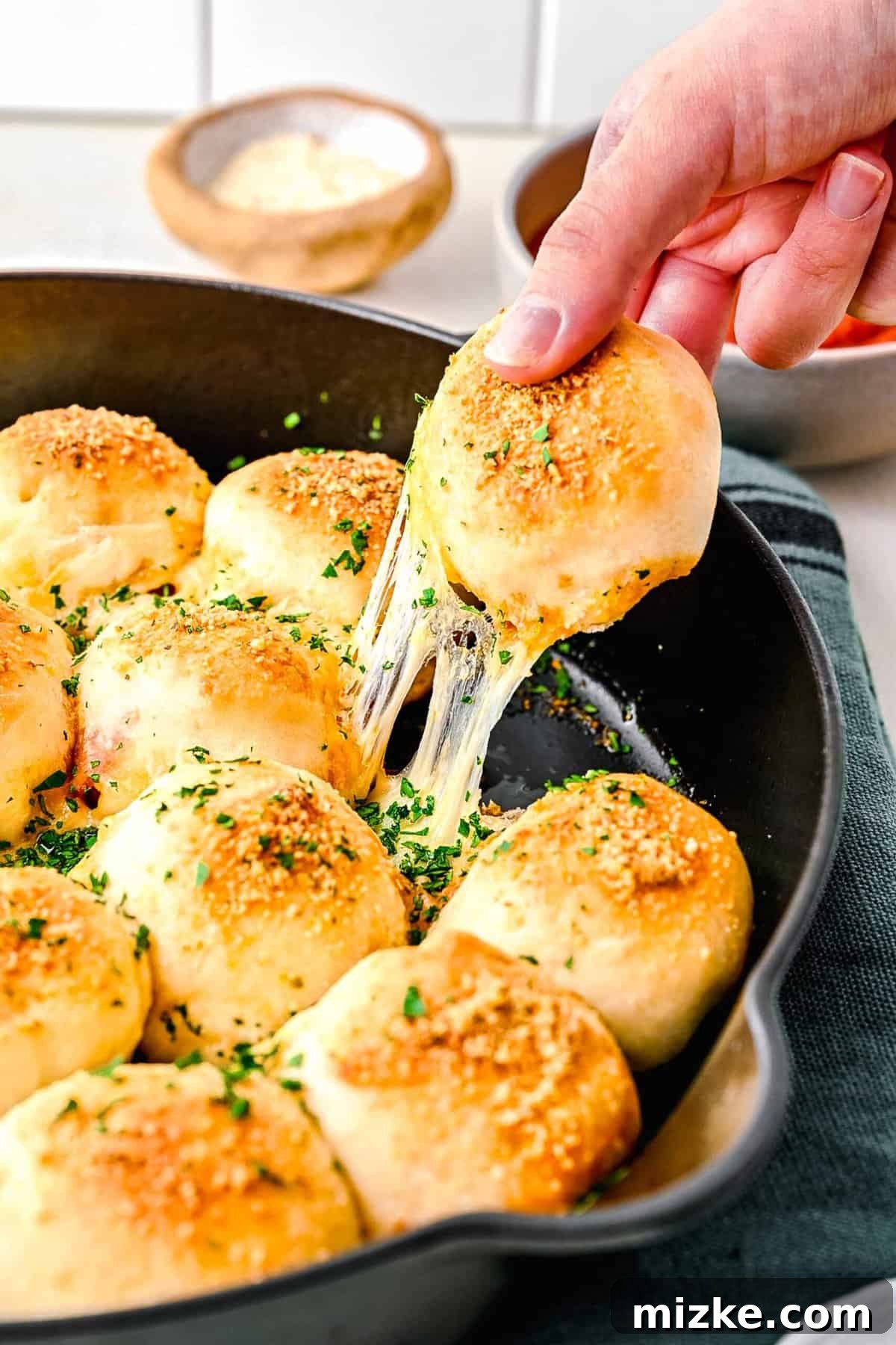 Golden-baked pizza bites, made with pizza dough wrapped around mozzarella and pepperoni, brushed with olive oil, and sprinkled with parmesan. A delicious skillet full of golden brown stuffed pepperoni pizza balls, with one ball having a bite taken out, revealing gooey melted cheese and pepperoni inside.