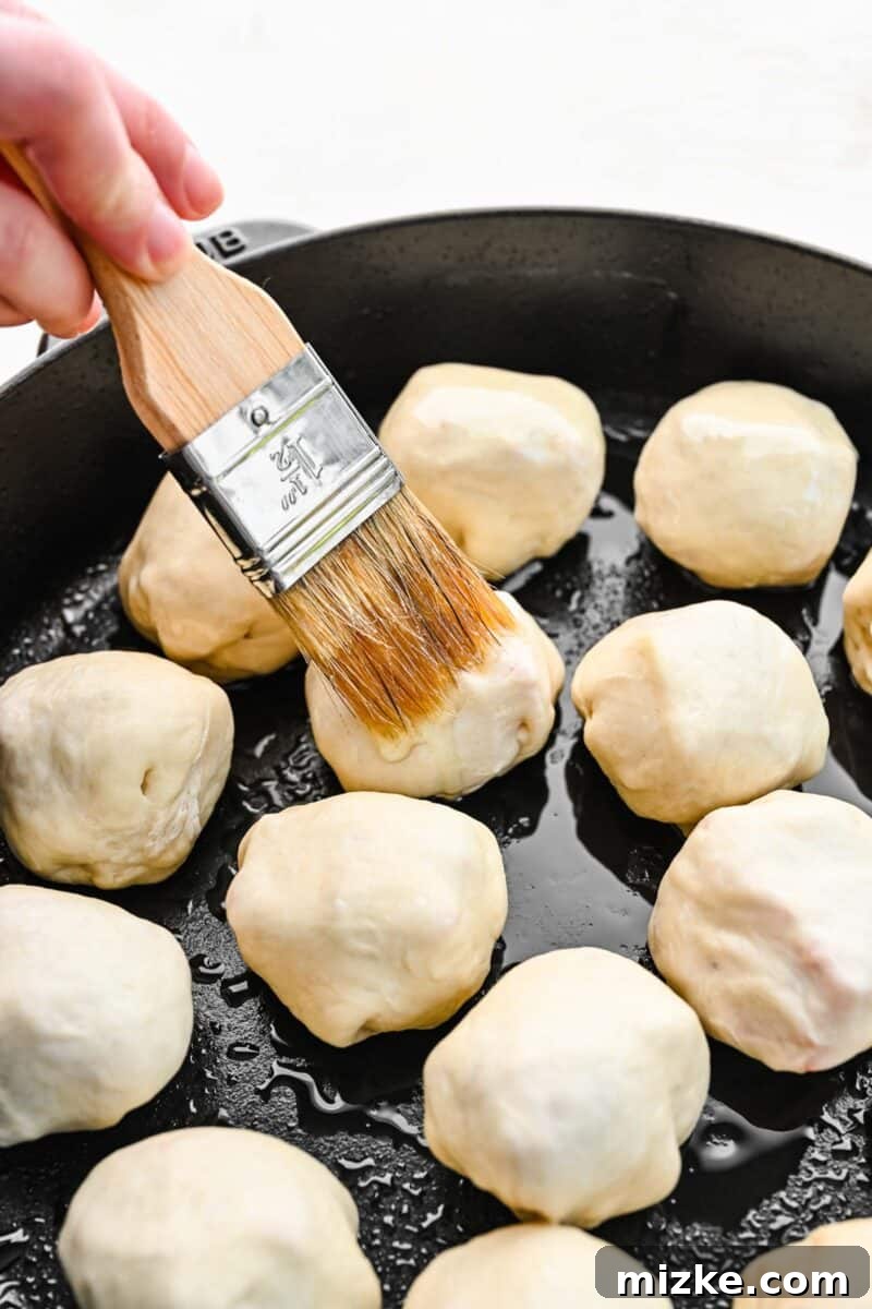 Brushing the pizza balls with olive oil before seasoning and baking. A pastry brush applying olive oil to the tops of the raw pizza balls in the skillet, ensuring even coating.