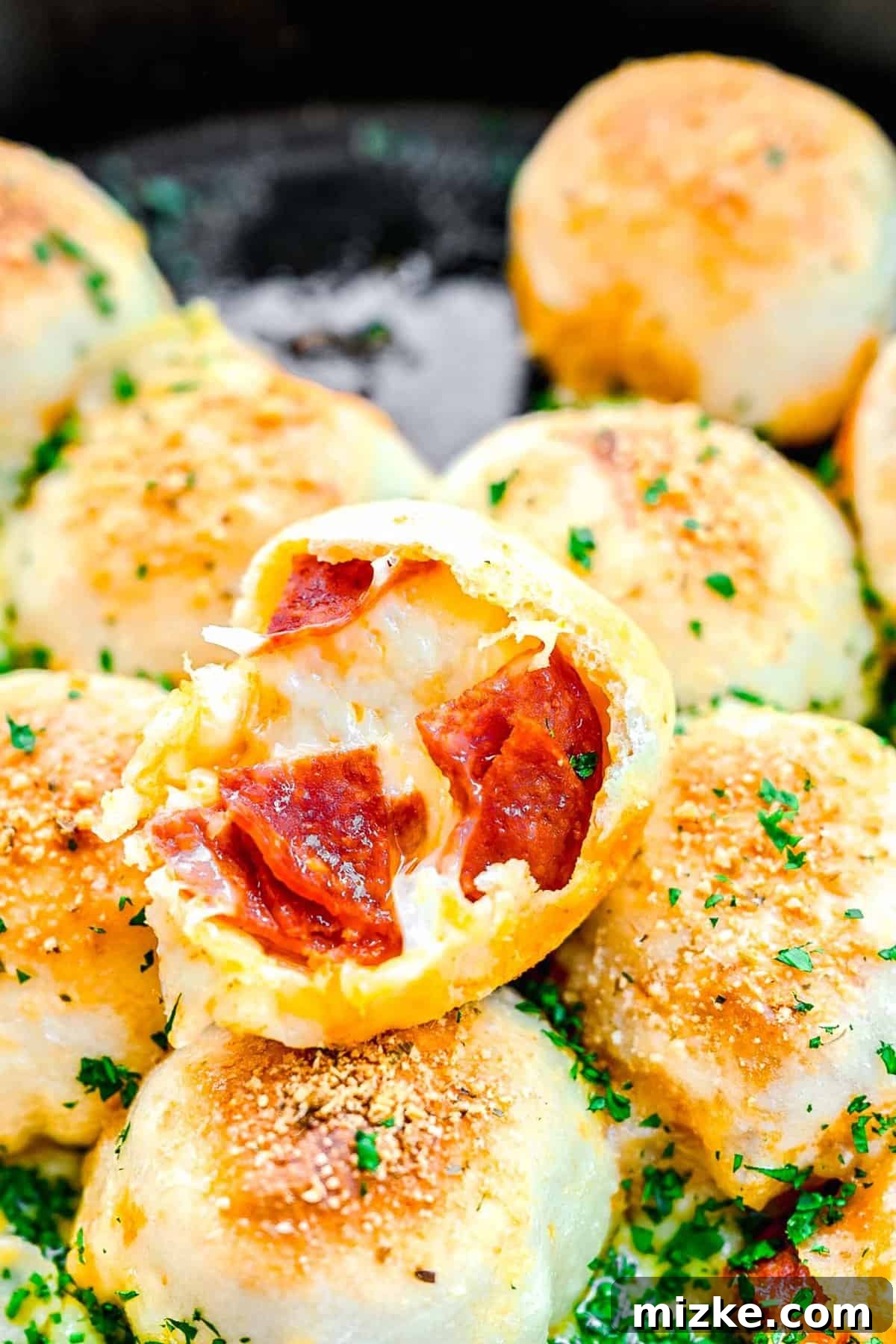 Freshly baked pizza bites, crafted from pizza dough encompassing mozzarella and pepperoni, then brushed with olive oil and parmesan. An overhead shot of a cast iron skillet brimming with freshly baked stuffed pepperoni pizza balls, one with a delightful bite missing, showcasing its cheesy interior.