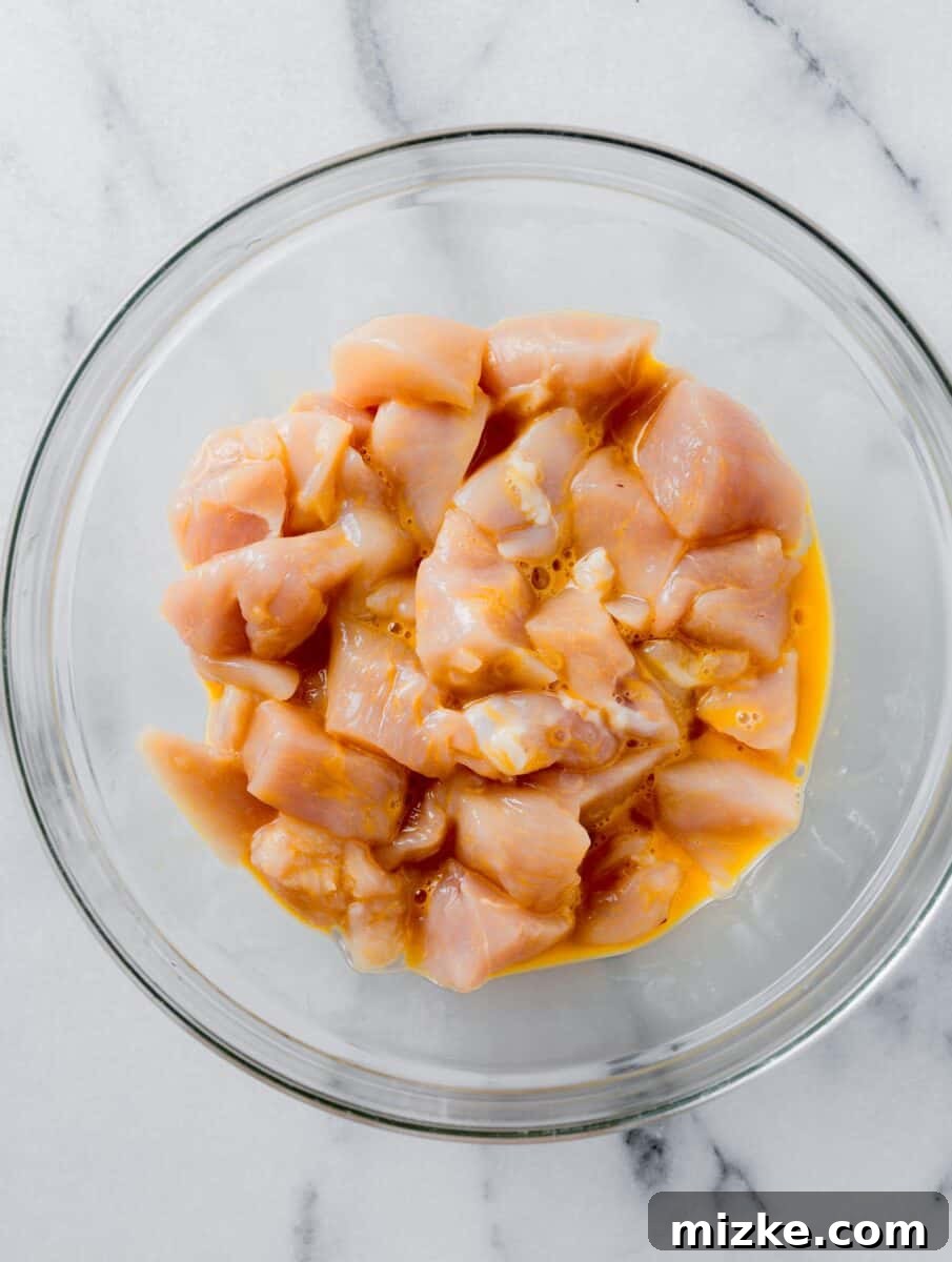 Raw chicken cubes tossed in an egg mixture in a clear bowl, ready for breading