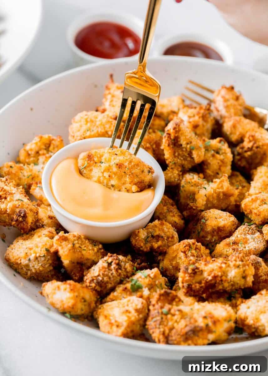 An air-fried chicken nugget being dipped with a fork into a bowl of dipping sauce, with other nuggets on a plate