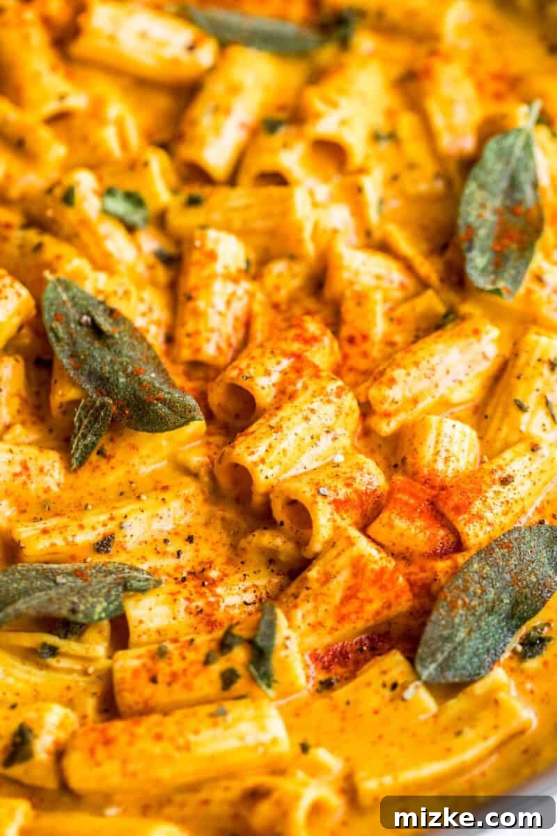 Velvet Vegan Pumpkin Pasta 3 A closer view of rigatoni pasta generously coated in a golden, creamy vegan pumpkin sauce, adorned with vibrant green sage leaves, a dusting of paprika, and a generous crack of black pepper, showcasing its inviting texture.
