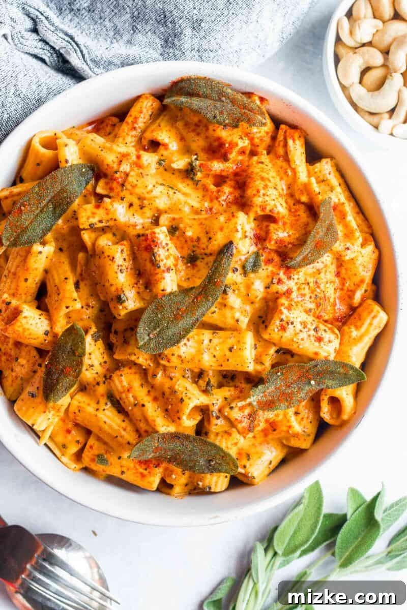 Velvet Vegan Pumpkin Pasta 5 A beautifully composed shot of rigatoni pasta in a creamy vegan pumpkin sauce, topped with fresh sage, paprika, and black pepper, presented in a white bowl alongside fresh sage sprigs and a metal fork, ready to be enjoyed.