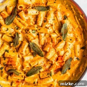 Rigatoni pasta in a creamy vegan pumpkin sauce, served in a large metal skillet and beautifully garnished with fresh sage leaves, a sprinkle of vibrant paprika, and freshly ground black pepper.