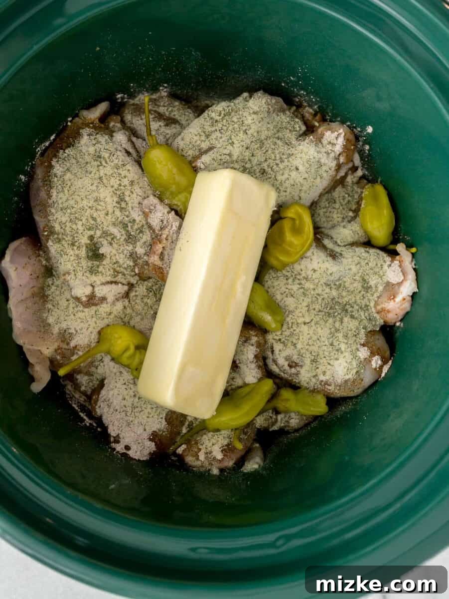 Zesty Mississippi Chicken 6 Chicken in a green ceramic slow cooker insert with au jus gravy mix and ranch seasoning sprinkled on top along with pepperoncinis and a stick of butter