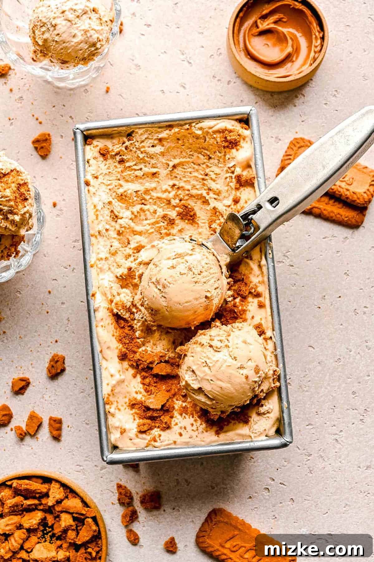 No-churn Biscoff ice cream in a loaf pan, ready to serve. An overhead view of an ice cream scoop delicately lifting creamy Biscoff ice cream from a loaf pan, surrounded by whole Biscoff cookies and a jar of cookie spread.