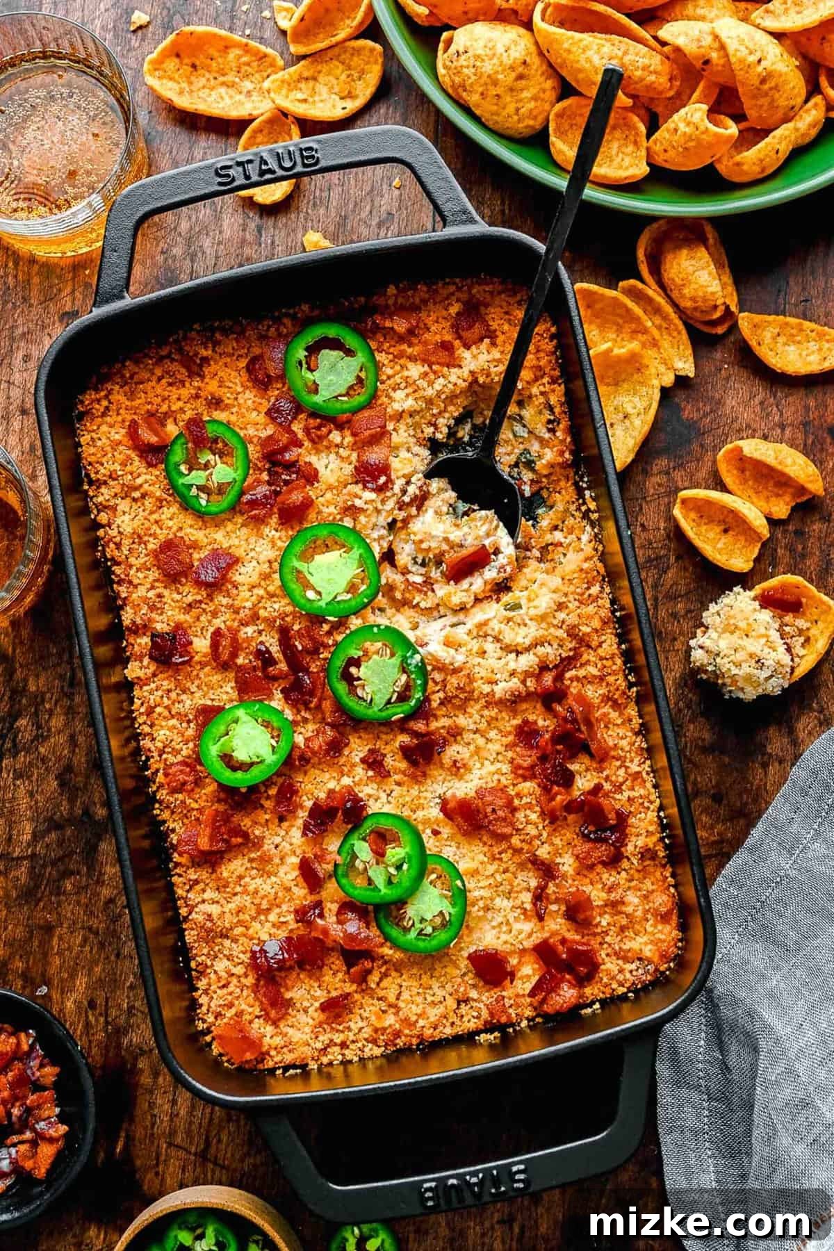 Overhead shot of a bubbly, golden-brown Jalapeño Popper Dip in a baking dish, with a scoop removed, revealing its creamy interior. A bowl of tortilla chips is nearby.