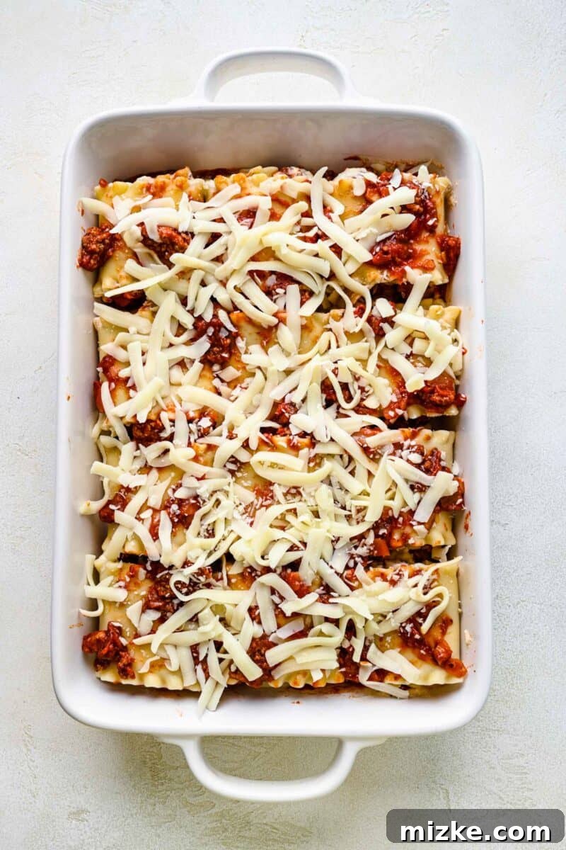Adding shredded mozzarella cheese to lasagna roll ups for a cheesy crust. Sprinkling generous amounts of shredded mozzarella cheese over lasagna roll ups in a baking pan, ready for the oven.