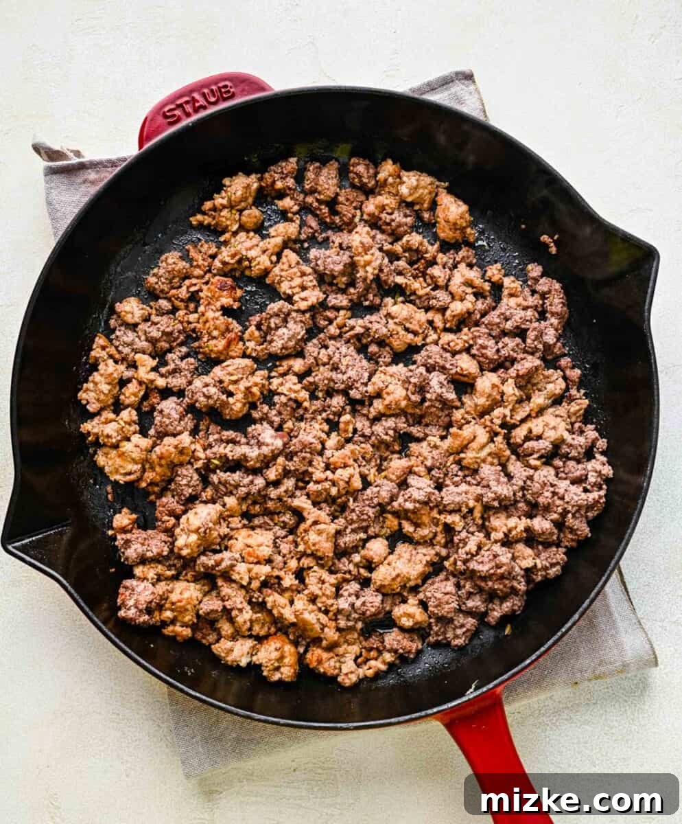 Searing ground beef and Italian sausage for the flavorful meat sauce. Close-up of ground beef and Italian sausage browning in a large skillet over medium heat, being broken apart with a wooden spoon.