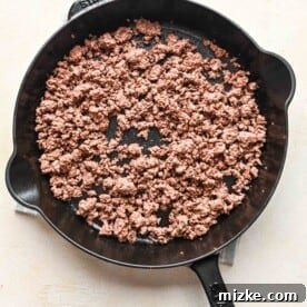 Cooked ground beef broken into smaller pieces in a cast iron skillet, seasoned and ready for the next step.