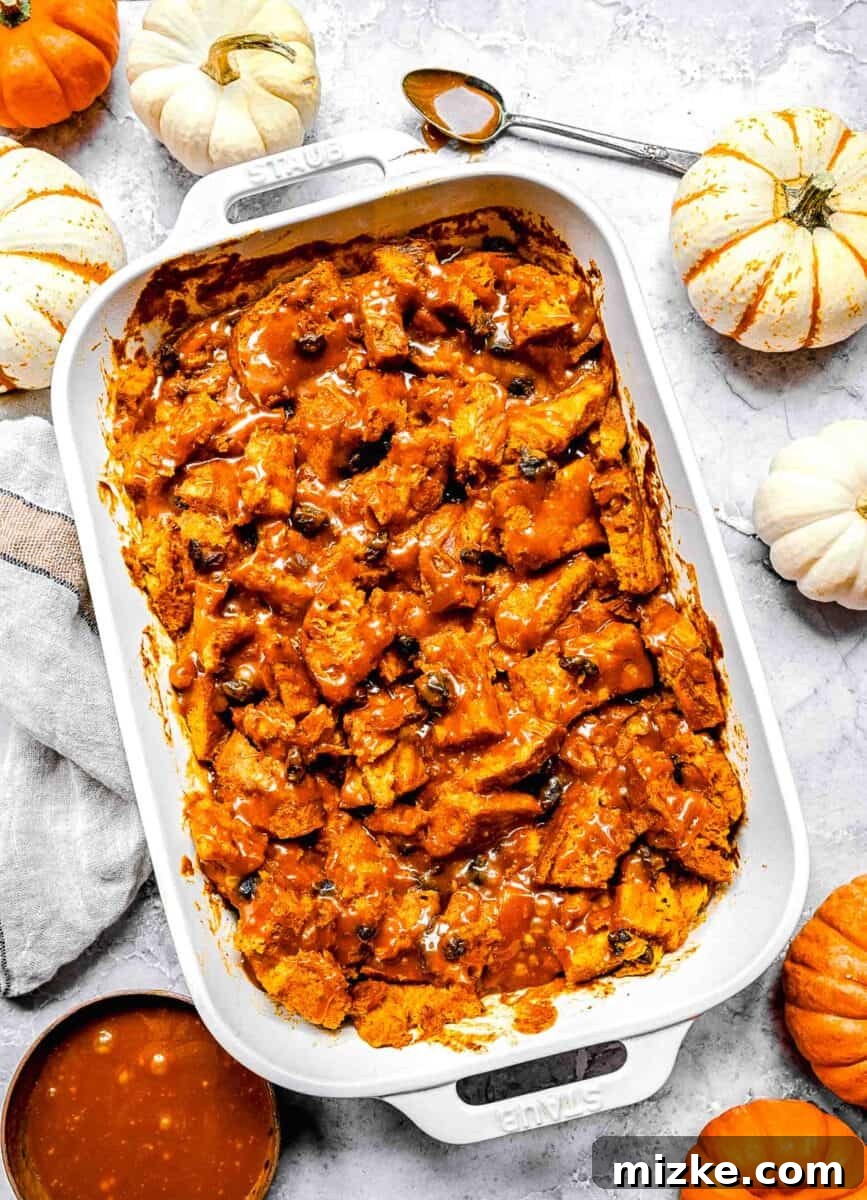 Golden-brown baked pumpkin bread pudding in a white casserole dish, generously drizzled with salted caramel sauce. Several small decorative pumpkins are arranged around the dish, enhancing the autumnal theme.